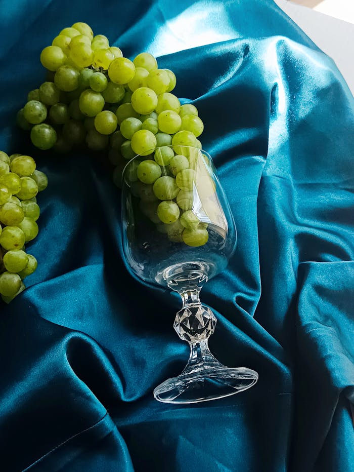 who-we-are A sophisticated display of green grapes with a wine glass on luxurious blue satin creates a vibrant and eye-catching composition.